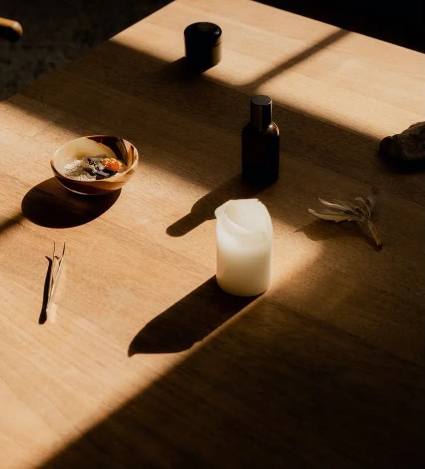 Objetos rituales sobre una mesa de madera bañada por la luz de la tarde: un cuenco, un frasco ámbar, una vela y un manojo de salvia.