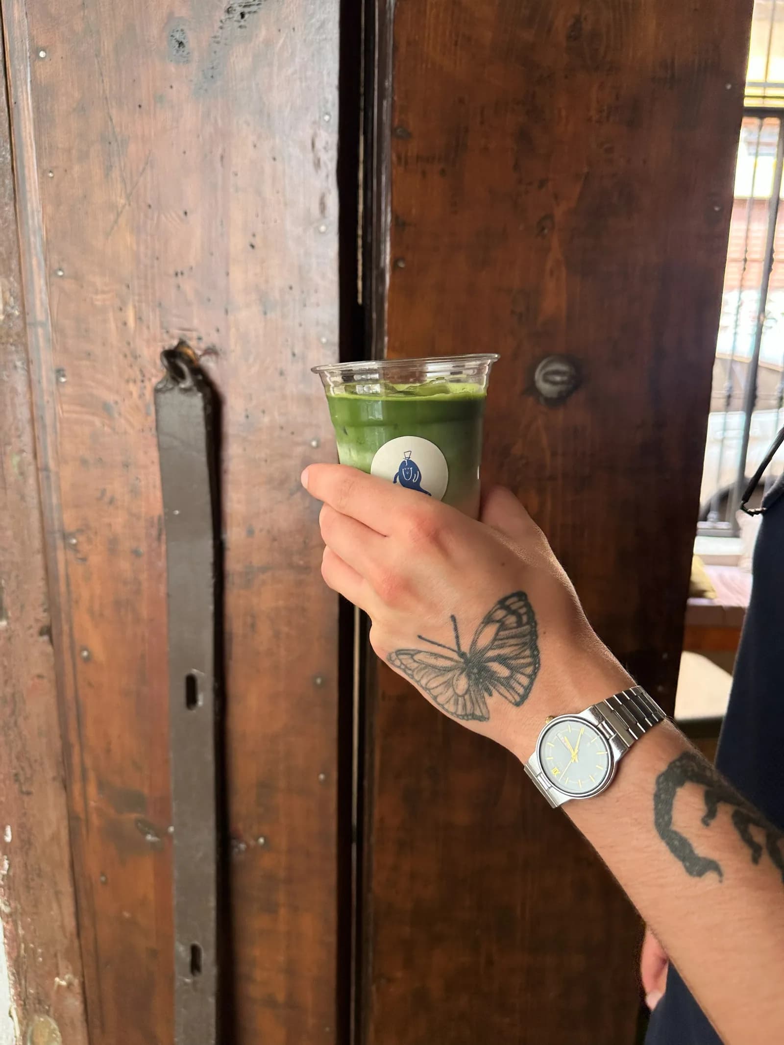 Una mano con un matcha para llevar junto a una puerta de madera.